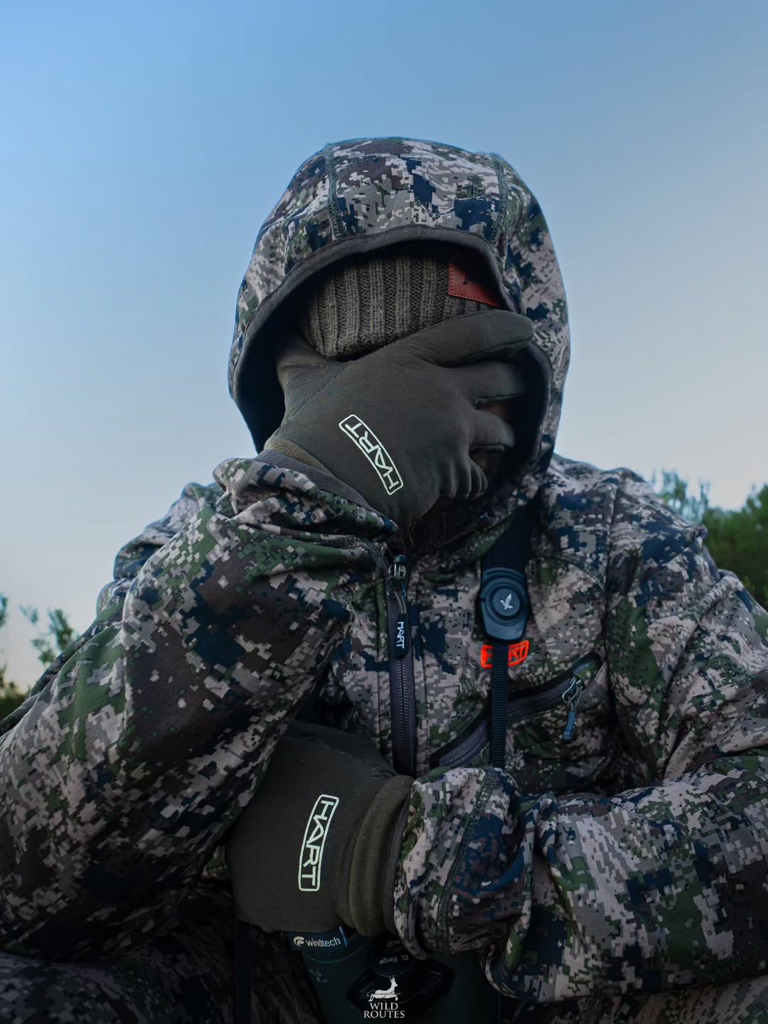 Chasseur en tenue camouflage Hart, capuche et jumelles, ambiance crépusculaire en forêt